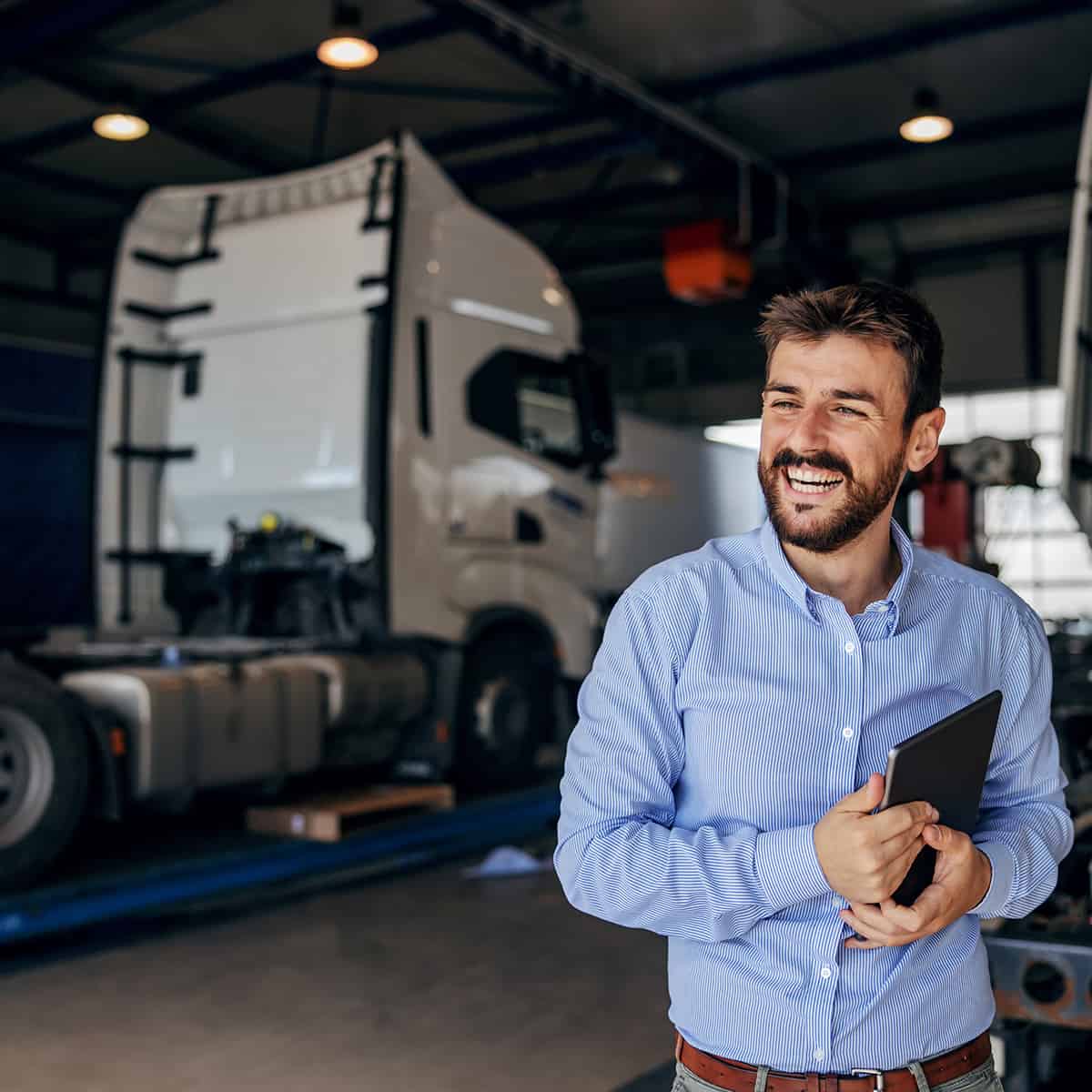 Smiling chief standing in auto park and holding tablet. In background are trucks. Firm for import and export.